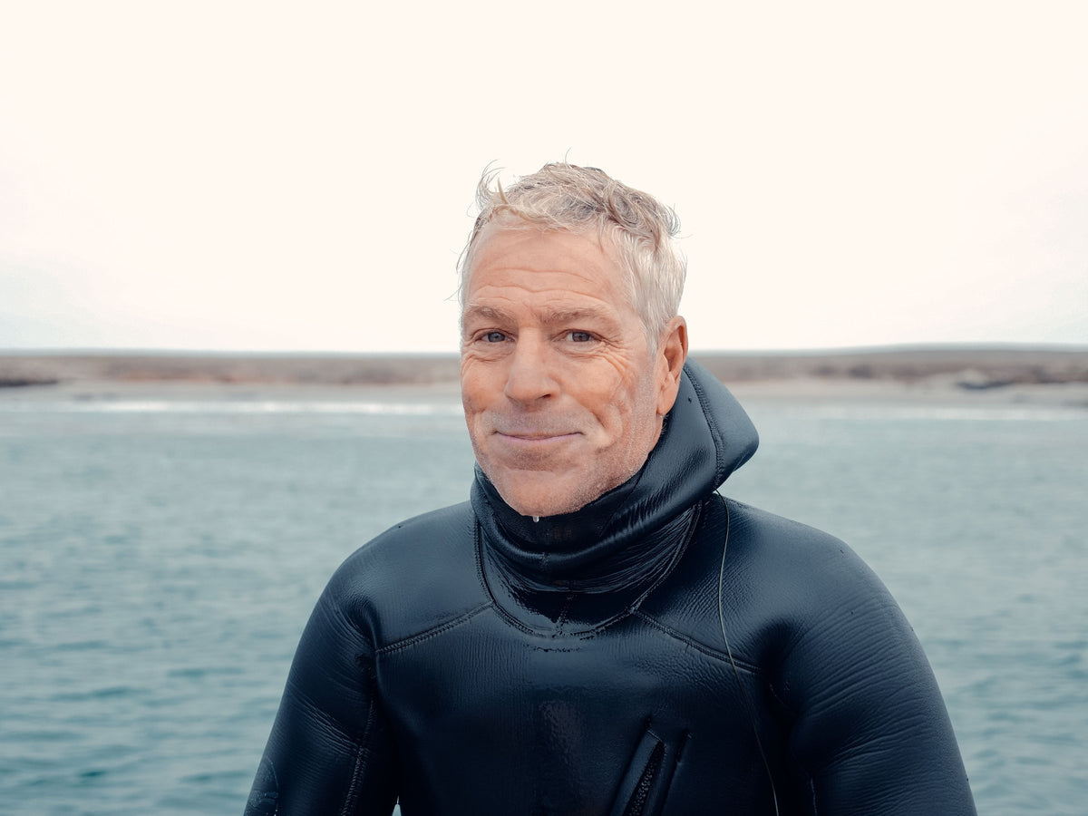 An older man in a hooded wetsuit standing beside the water before a dive