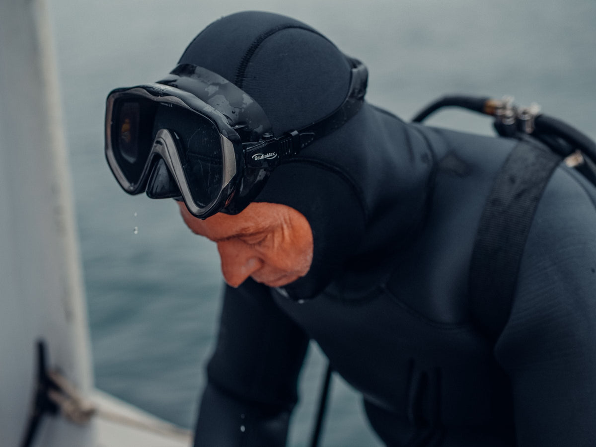 Older diver in a thick hooded neoprene suit and mask leaning over a boat side, grey overcast sea behind, water dripping off the mask