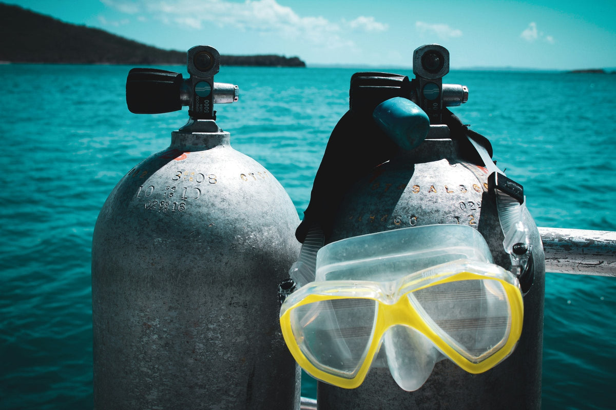 Two aluminium scuba cylinders with valves on a boat, a clear yellow dive mask hanging on one, turquoise sea behind, Fajardo, Puerto Rico