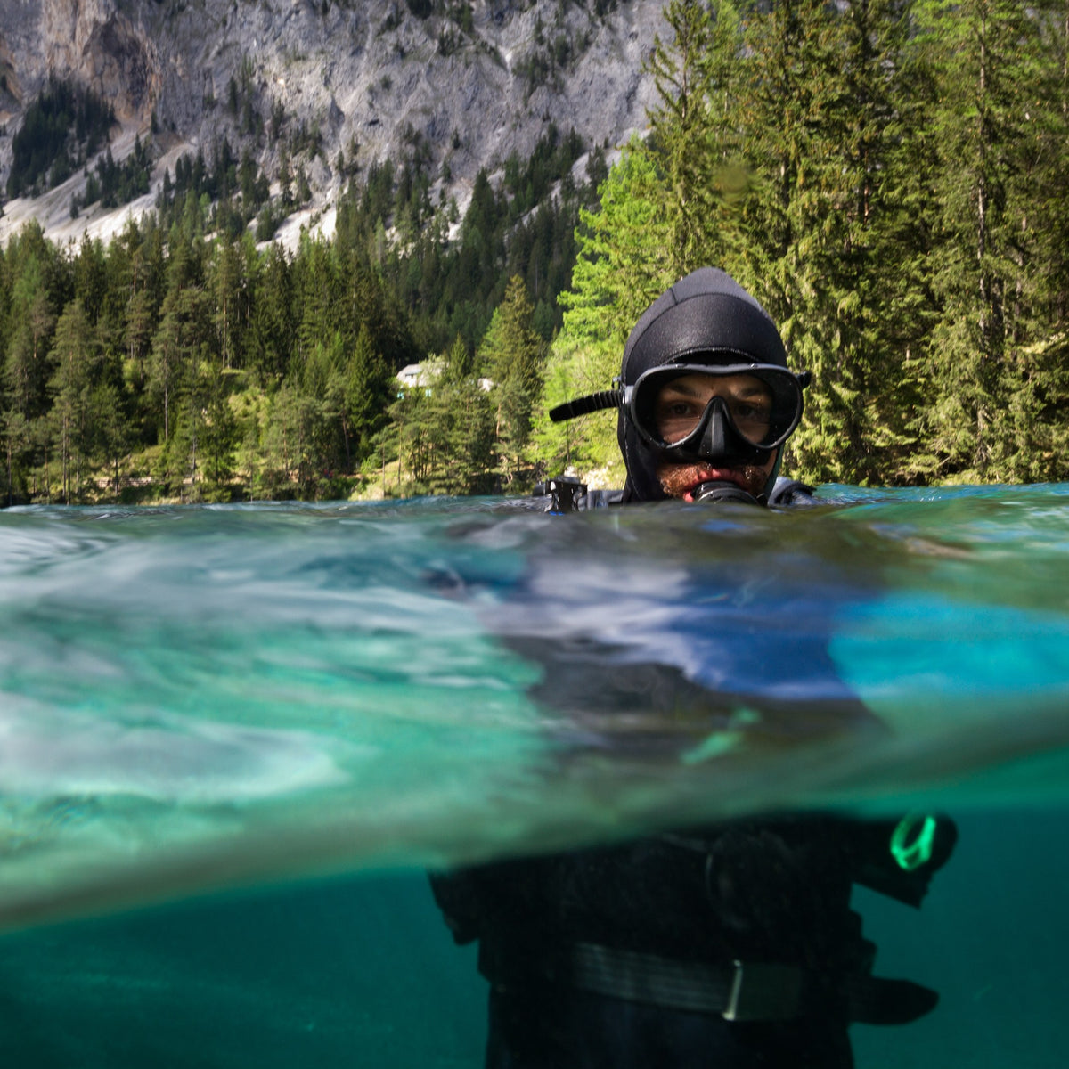 Diver surfacing in an alpine lake under forested cliffs
