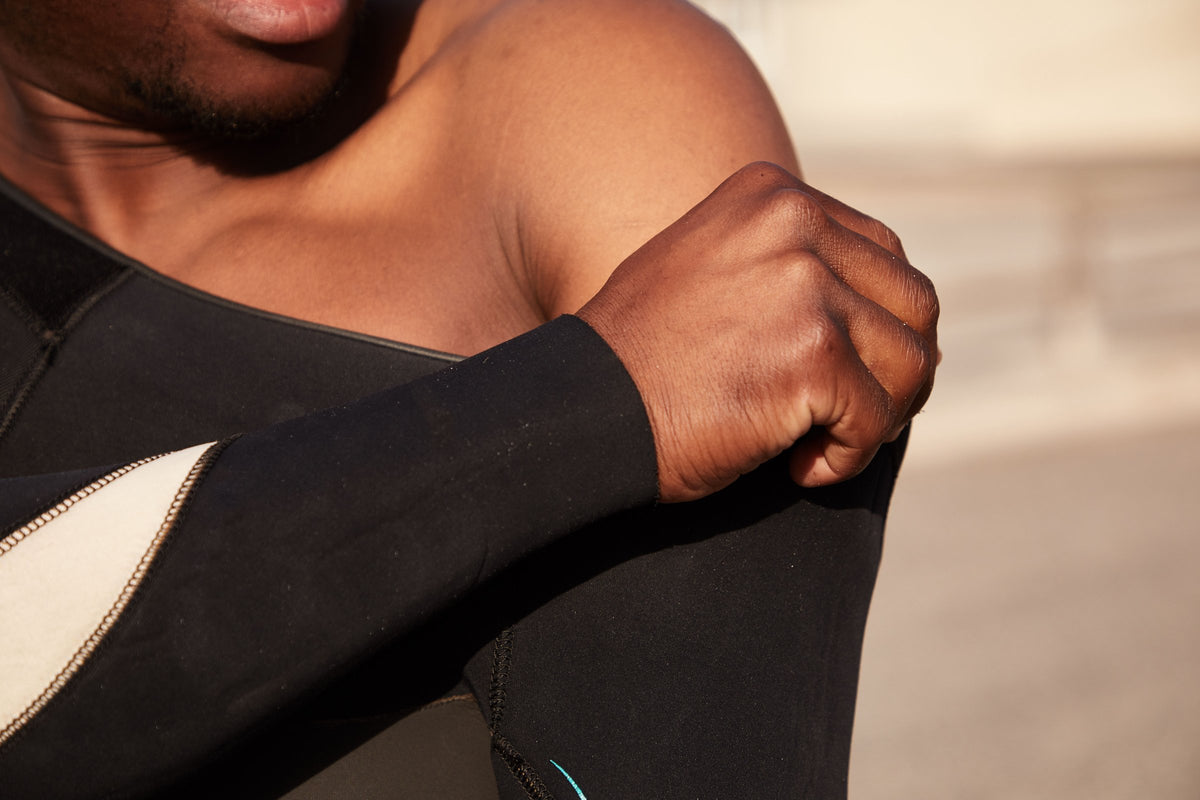 Close crop of a shoulder and arm pulling on a black neoprene wetsuit on a shoreline