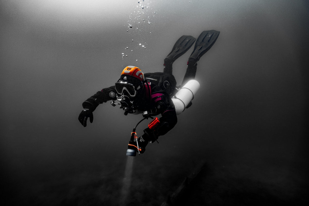 Sidemount diver with a helmet and torch in near-monochrome dark water, sparse bubbles