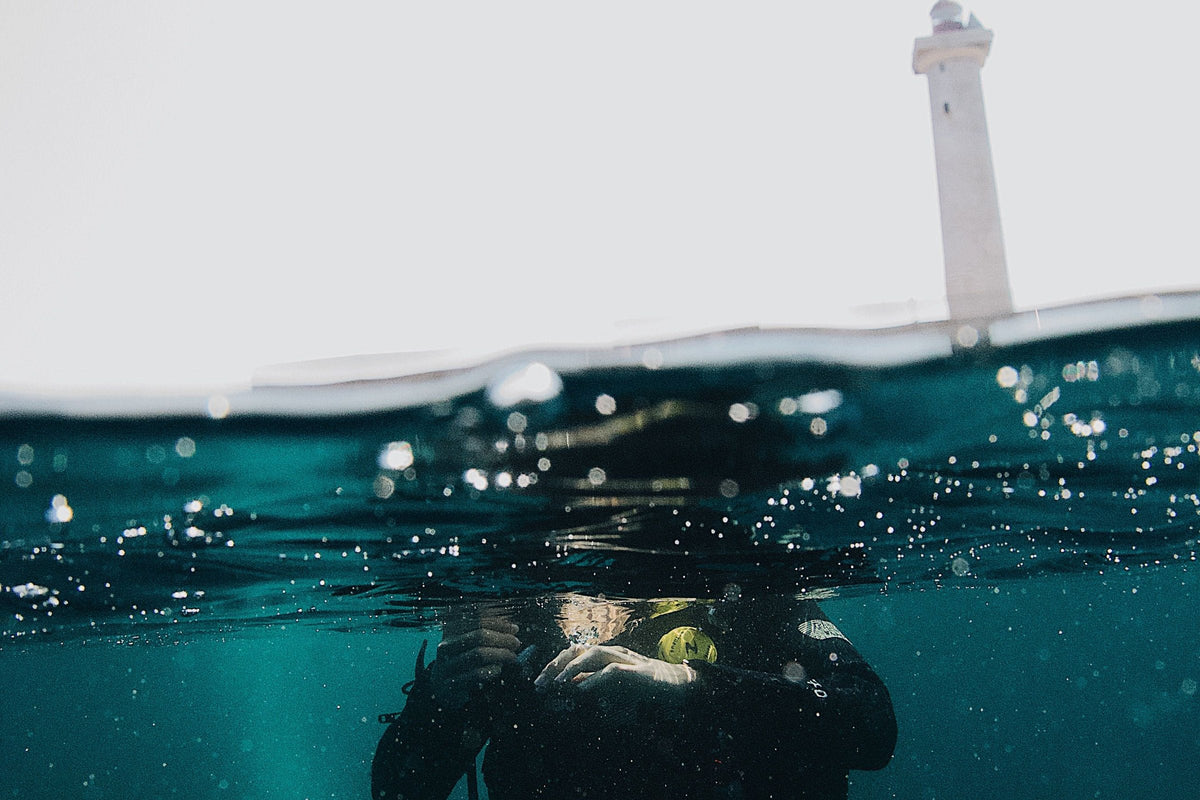A diver floating at the surface seen half above and half below the waterline, a lighthouse on the shore behind