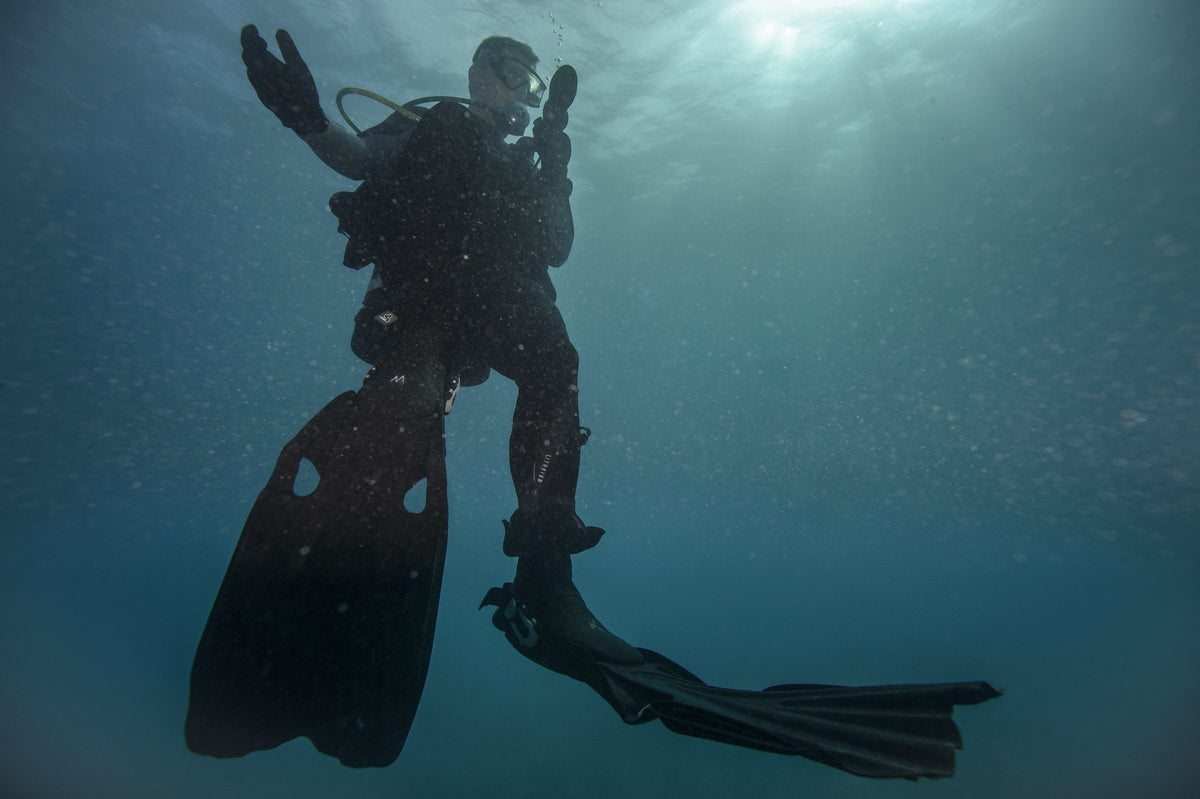 Silhouetted diver hanging head-up below a sunlit surface, reading a gauge console, long fins below