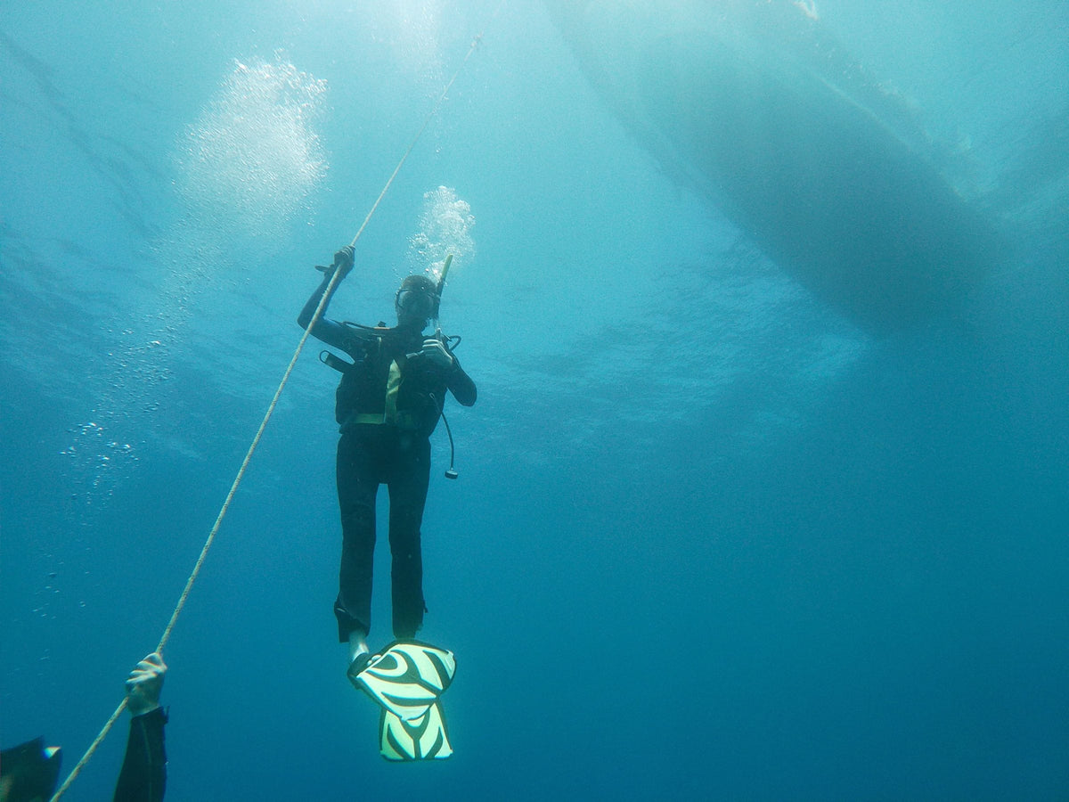 Diver holding an ascent line beneath a boat hull during a safety stop, bubbles rising
