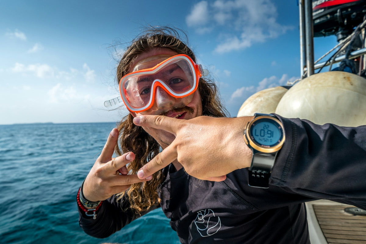 Diver on a boat deck holding a wrist dive computer up to the camera, sea and sky behind
