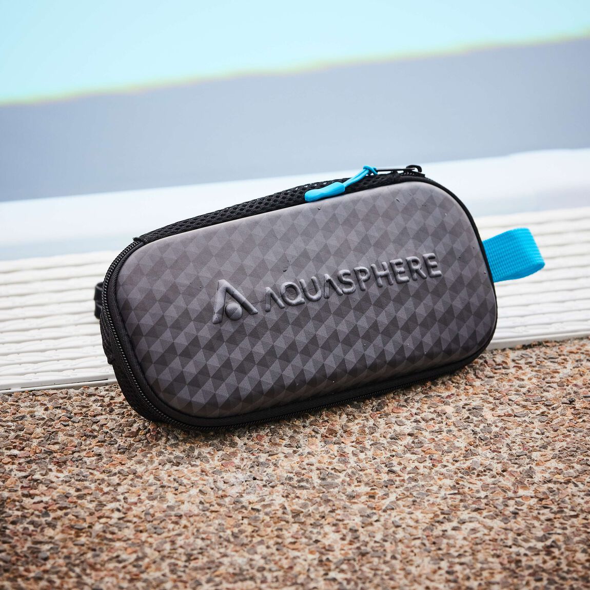 Closed Aquasphere mask case with its blue tab on a pool deck