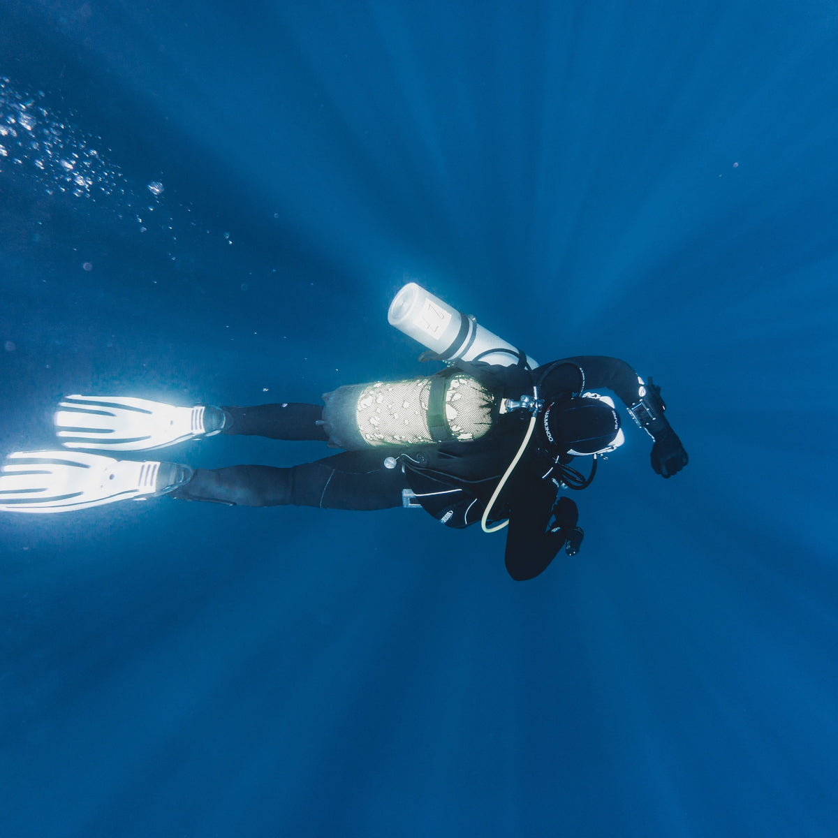 Solo diver carrying a slung redundant cylinder in open blue water under sun rays